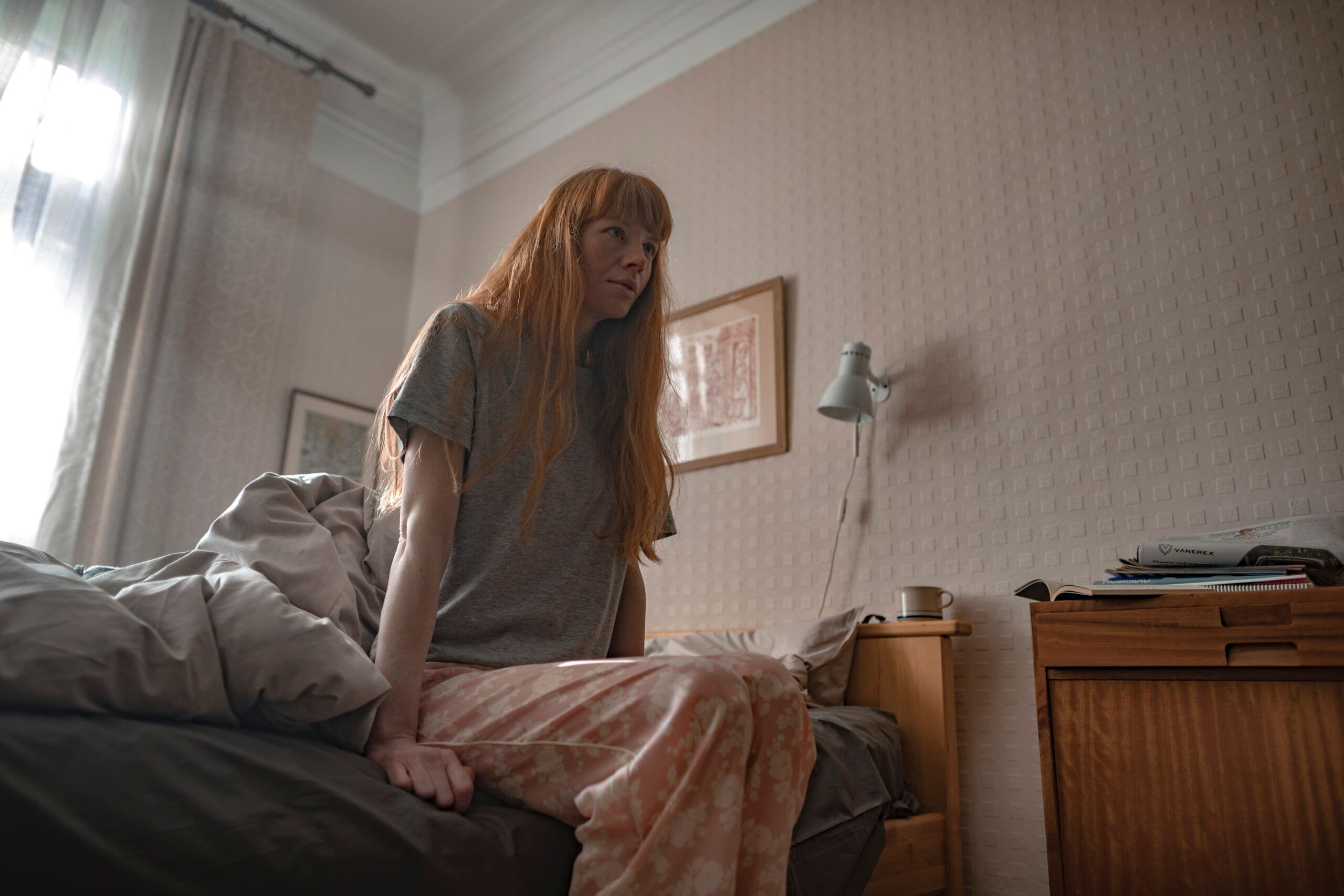 Inimene istub voodi serval magamistoas, taustal raamitud pildid ja öökapp.