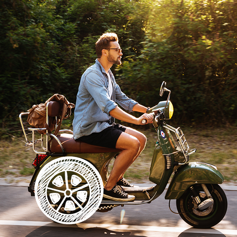 Inimene sõidab rohelise motorolleriga mööda teed, taustal rohelus; seljas helesinine teksasärk ja mustad lühikesed püksid, tagumisel pakiraamil pruun kott; tagumine ratas stiliseeritud valge kriidi või markerijoontega