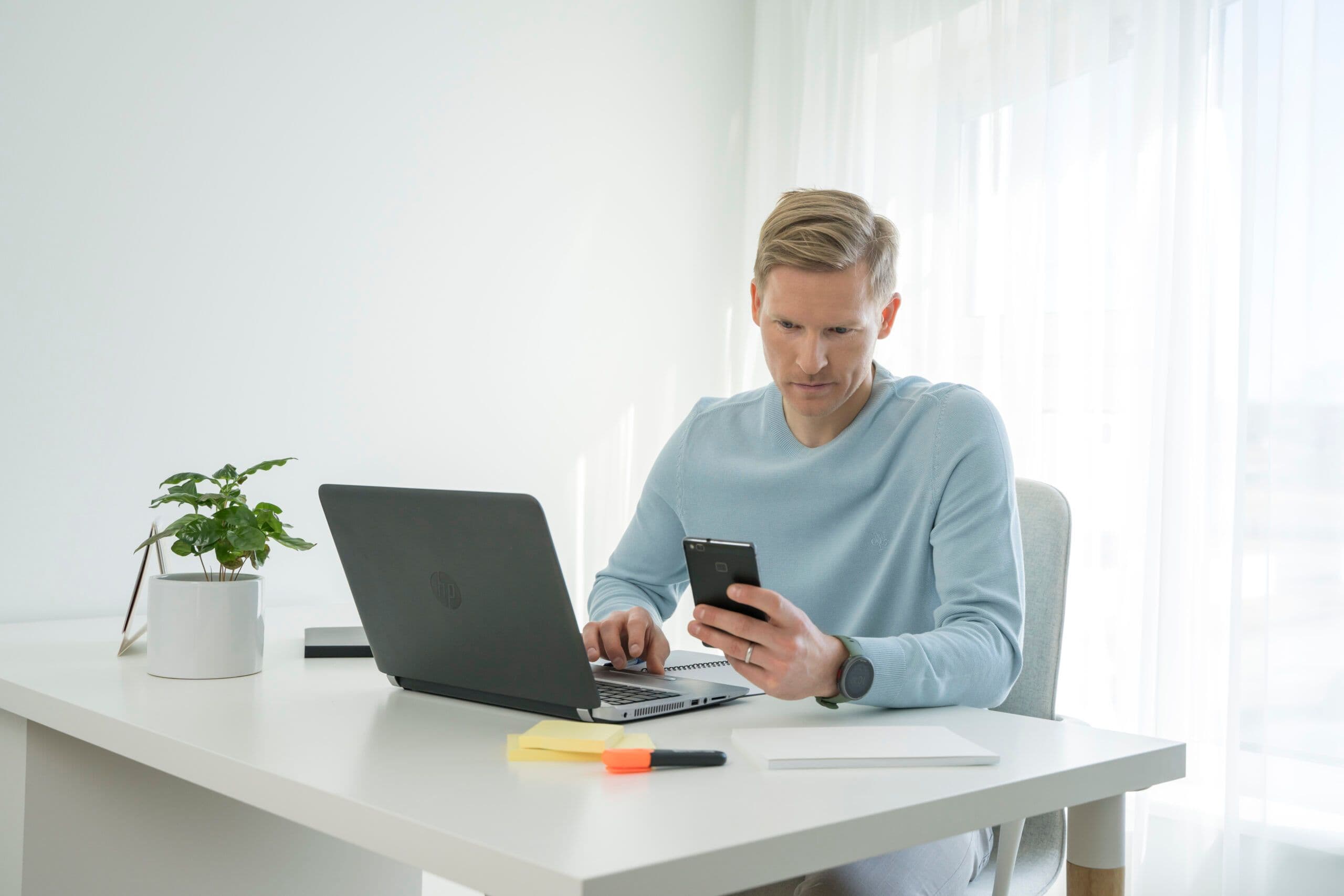 Inimene töötab sülearvutiga, hoides telefoni valgel laual