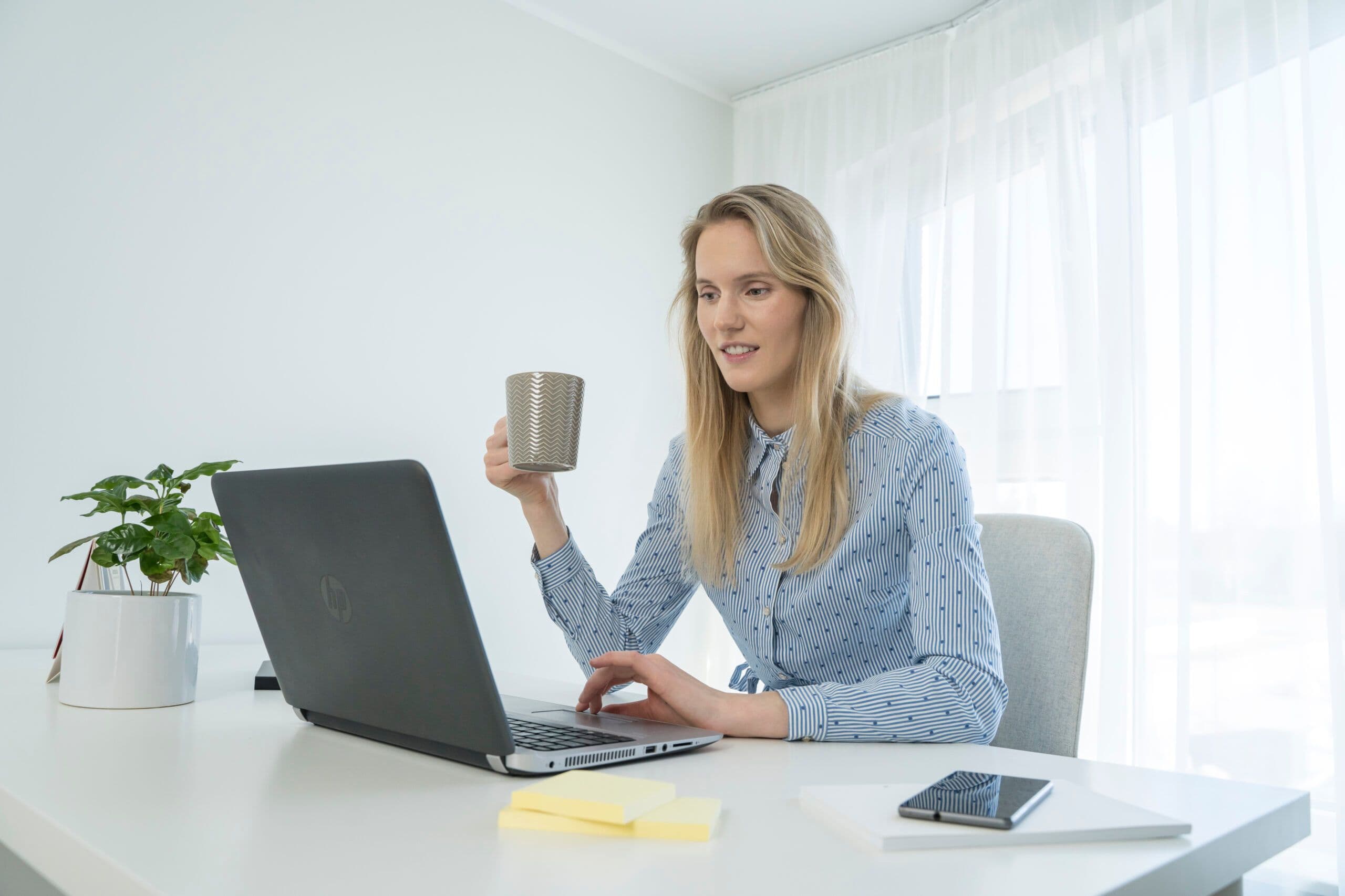 Inimene istub laua taga, hoiab ühes käes kohvitassi ja töötab teise käega sülearvutiga. Laual on nutitelefon, kalkulaator, kleepmärkmed ja potitaim. Taustal on valged kardinad ja päevavalgus paistab aknast sisse.