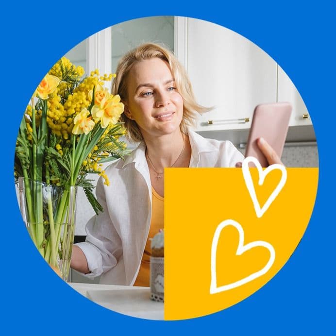 Inimene udustatud näoga hoiab nutitelefoni, istudes laua taga, millel on kollaste lillede vaas; taustal on köögikapid