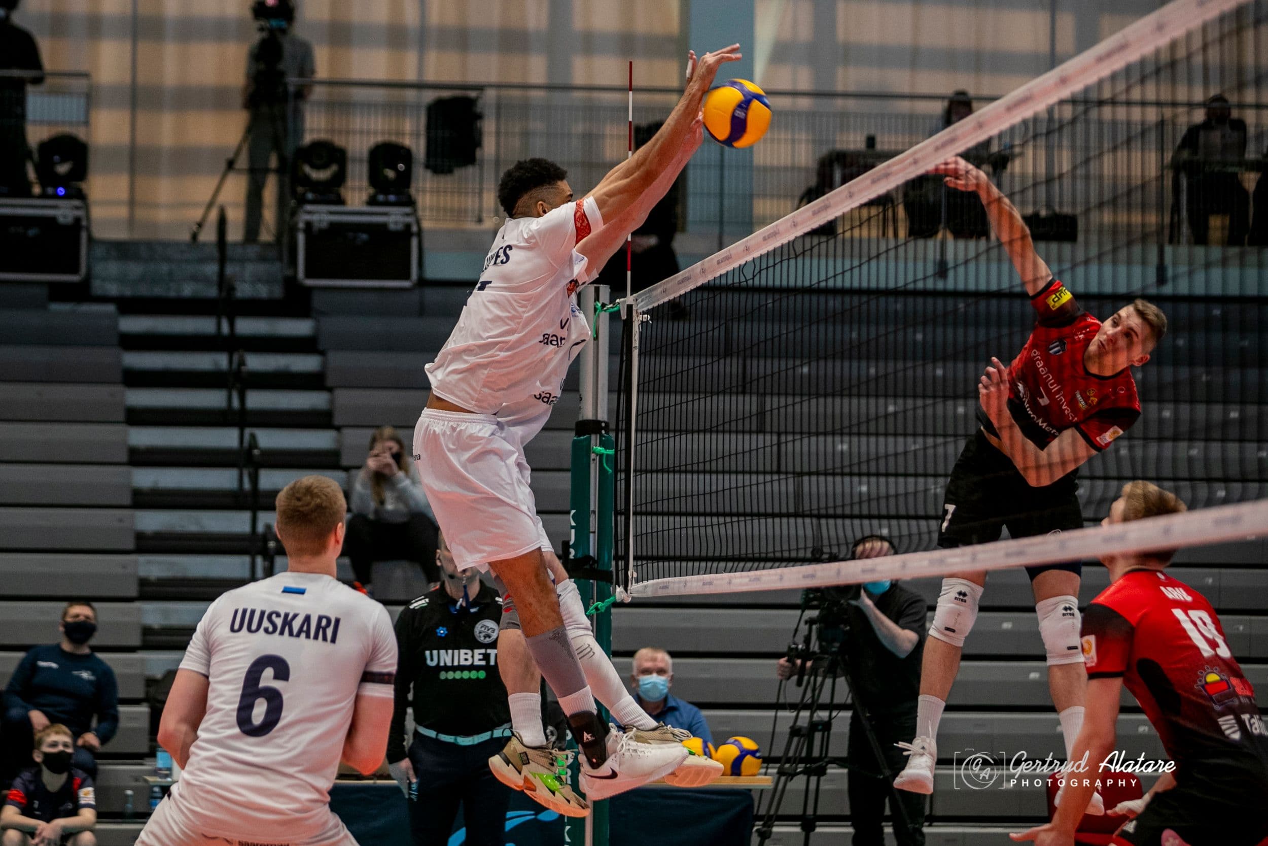 Sisehallis toimuv võrkpallimäng: valges vormis mängija hüppab, et blokeerida punases vormis mängija rünnakut. Taustal on näha mängija nimega 'UUSKARI' ja numbriga 6, teisi mängijaid ning pealtvaatajaid.