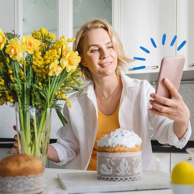 Inimene udustatud näoga hoiab nutitelefoni, istudes laua taga, millel on kollaste lillede vaas; taustal on köögikapid