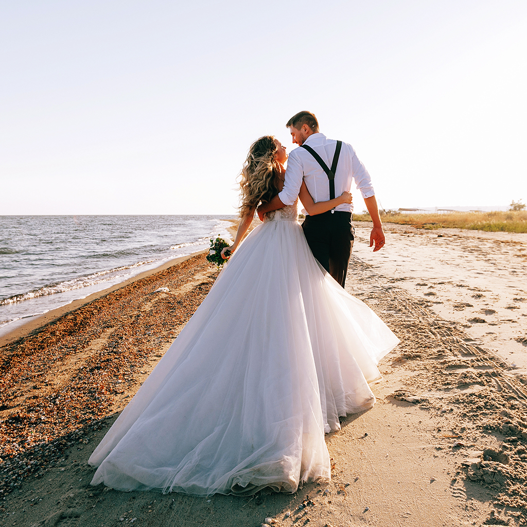 Noorpaar jalutab rannal, pruut valges kleidis ja peigmees traksidega särgis