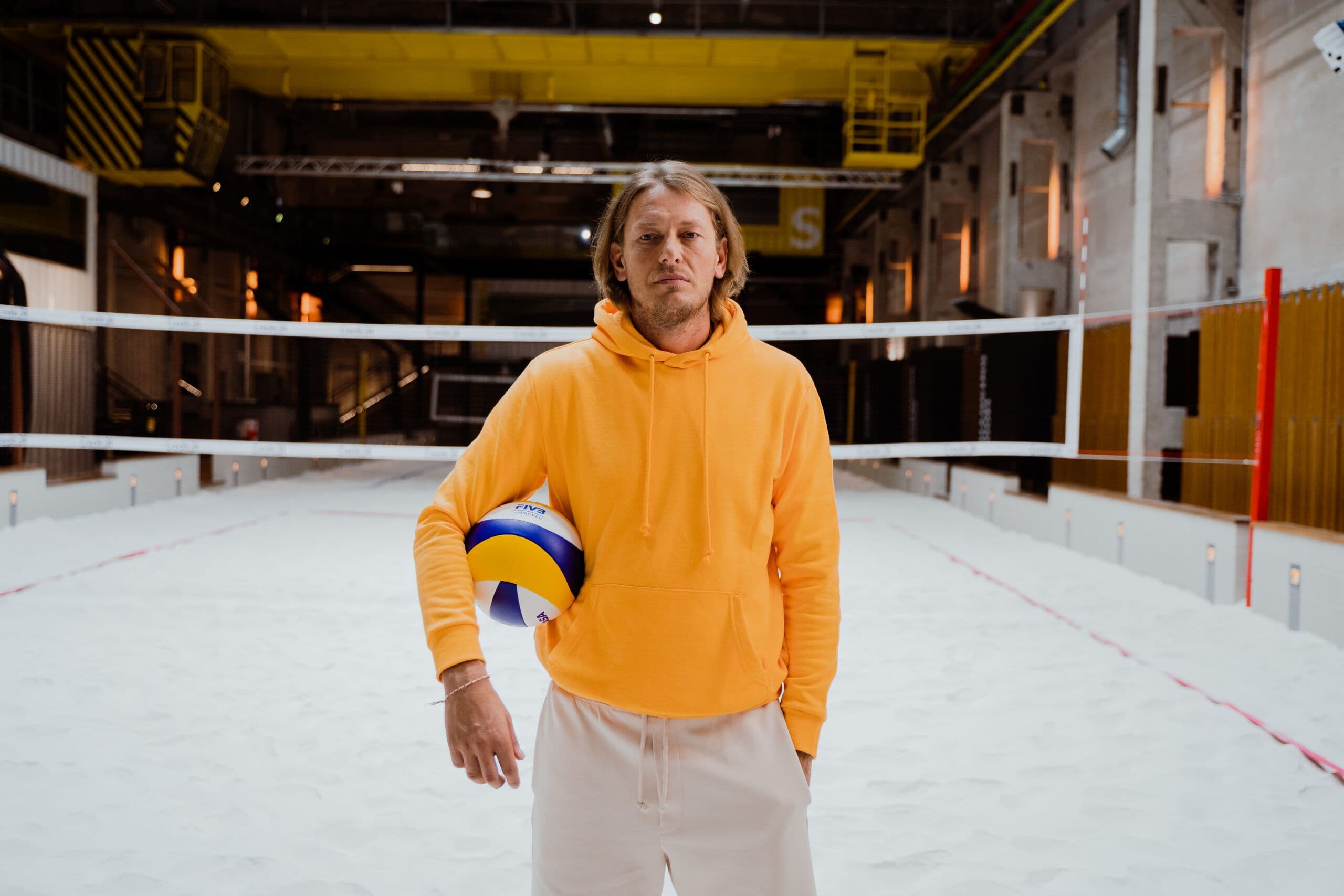 Inimene oranžis pusas seisab sise-rannavõrkpalliväljakul, pall kaenlas.