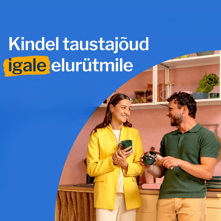 Inimene udustatud näoga hoiab nutitelefoni, istudes laua taga, millel on kollaste lillede vaas; taustal on köögikapid