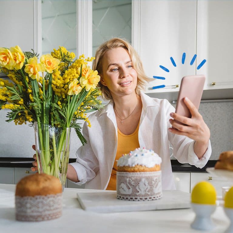 Inimene udustatud näoga hoiab nutitelefoni, istudes laua taga, millel on kollaste lillede vaas; taustal on köögikapid