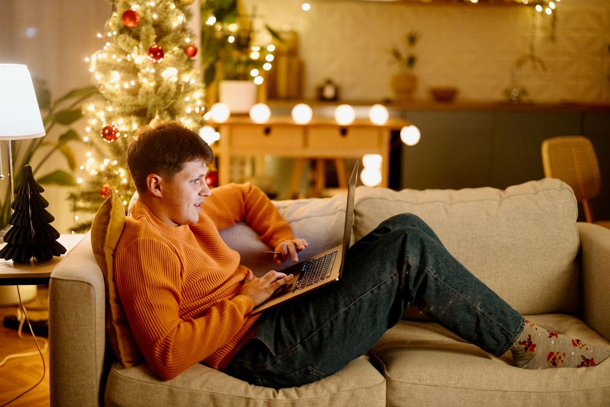 Inimene oranžis kampsunis istub diivanil ja kasutab sülearvutit, taustal jõulupuu